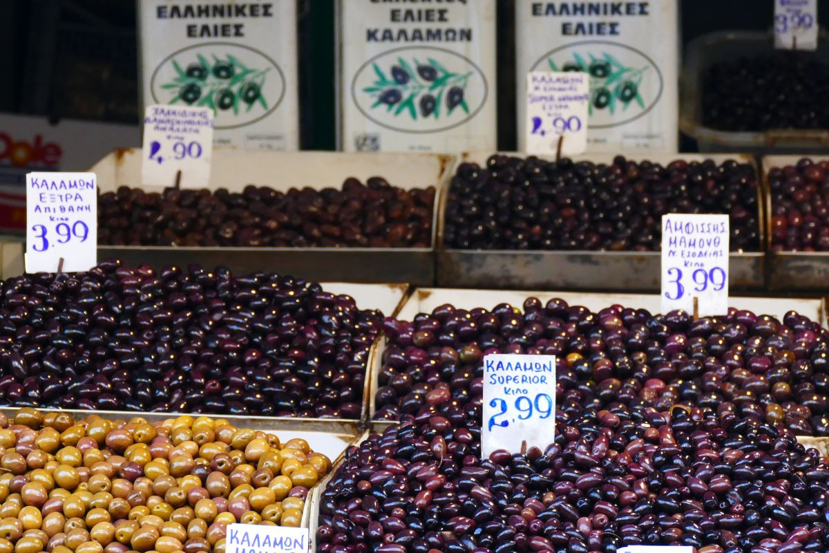 A variety of Greek olives, including Kalamon and green olives, displayed in bins at a market, with price tags visible and containers labeled in Greek.